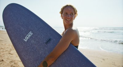Eine Frau steht am Strand, ein Surfbrett unter dem Arm.